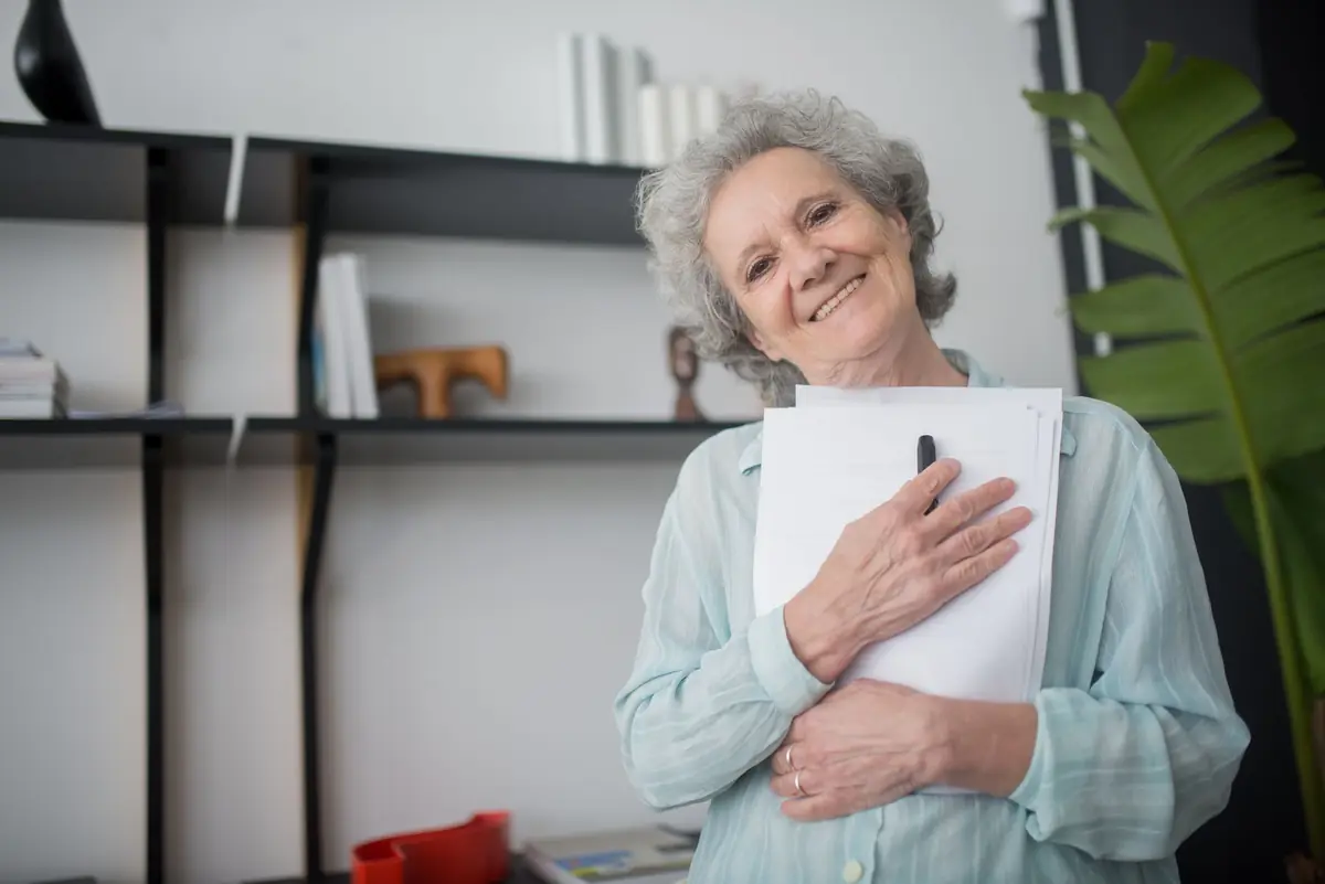 Senhora sorrindo em casa, segurando documentos junto ao peito